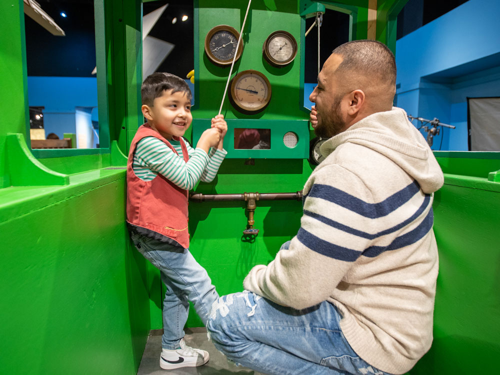A smiling child is pulling a rope to ring a bell inside the Dinosaur Train engine. An adult is kneeling beside the child.